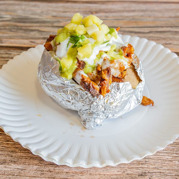 A foil-wrapped baked potato topped with seasoned meat, white sauce, green sauce, and pineapple chunks.