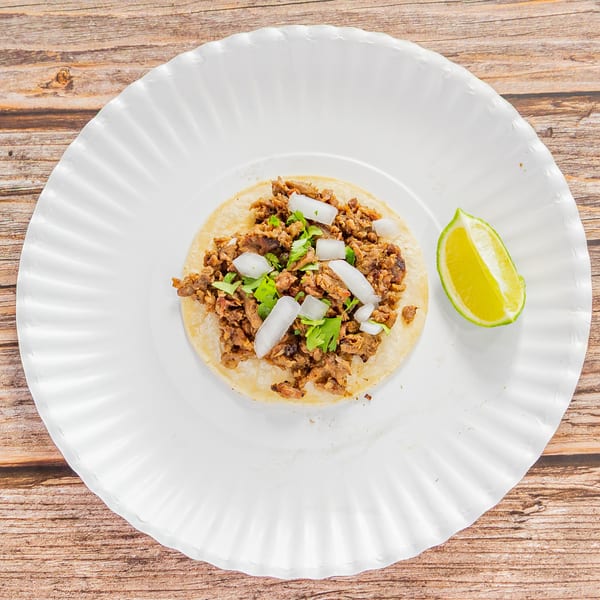 A single Tap N Taco carne asada taco with onions, cilantro, and a lime wedge on a white paper plate.
