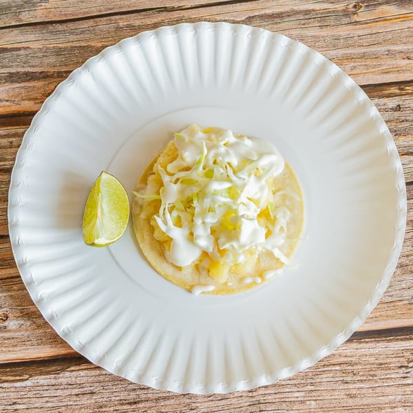 A Tap N Taco fish taco with cabbage, crema, and a lime wedge on a white paper plate.