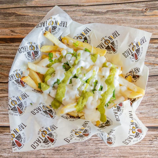 Tap N Taco's french fries topped with white and green sauces, and cilantro, on a wooden table.