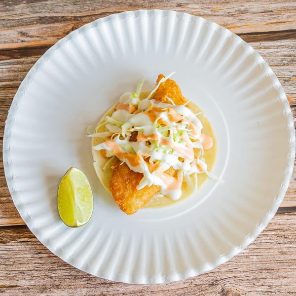 Crispy fish taco with shredded cabbage, white sauce, and orange sauce, served with a lime wedge.