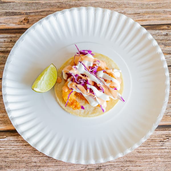 A Tap N Taco shrimp taco with purple cabbage, white and orange sauce, and a lime wedge.